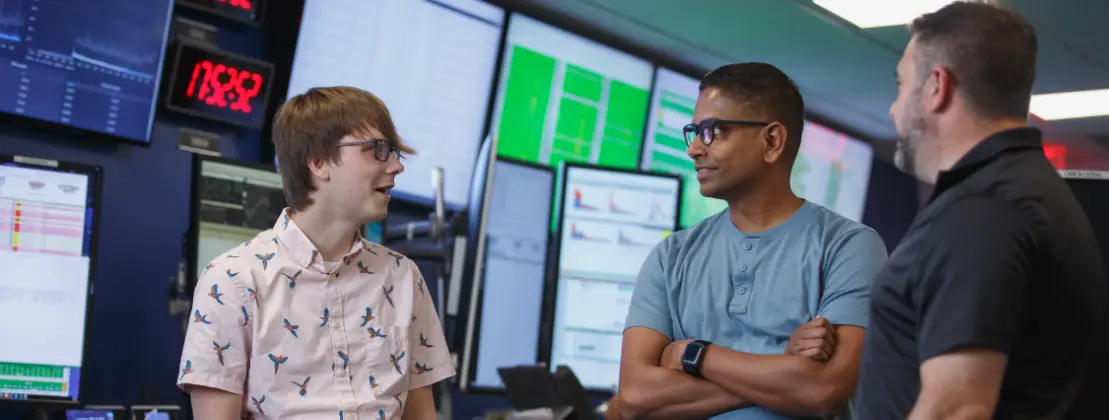 Three people standing in front of trading desk monitors